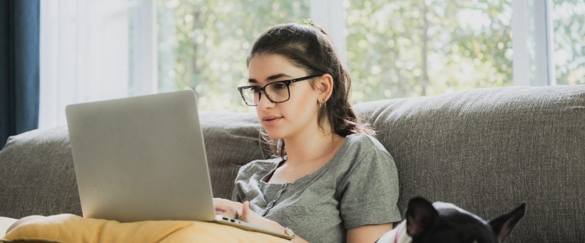 Funny and cheerful girl watching comedy movie on laptop with spectacles and pet dog while relaxing at home sitting on couch