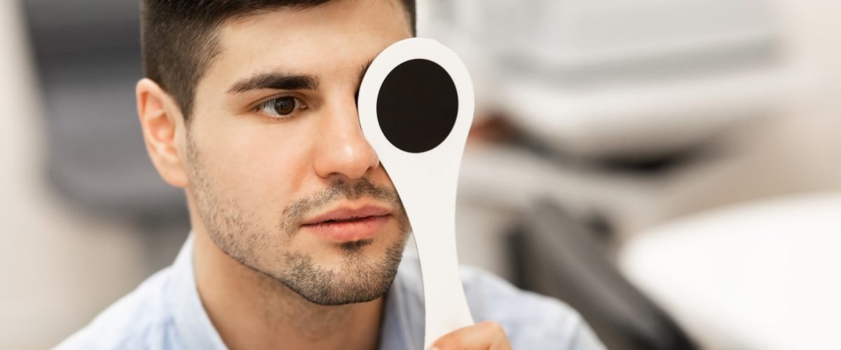 Poor Vision Concept. Closeup portrait of serious man reviewing eyesight, closing eye with occluder, looking at test chart. Male client doing test in clinic. Banner, panorama, blurred background